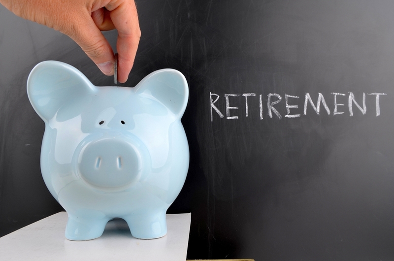 Floating fingers depositing a coin into a blue piggy bank with the word 'retirement' written in chalk on a blackboard behind it.